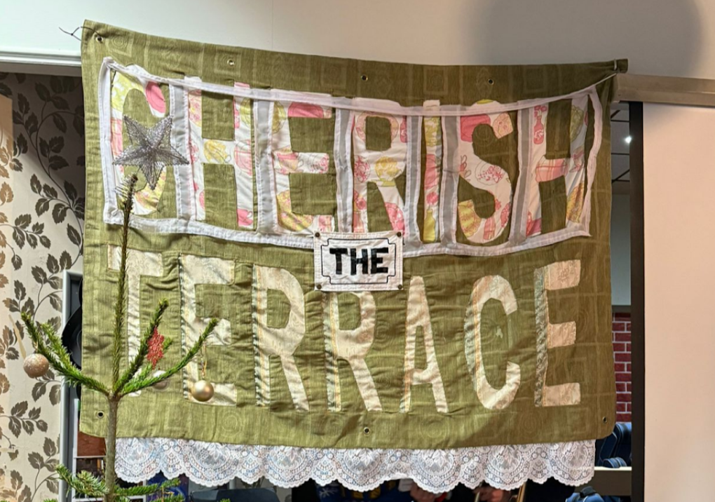 A photograph of a handmade banner. A large green textured blanket has lace along the bottom side and in cut-out materials ‘cherish the terrace’ in full capital letters is spelled. Each word is on a new line, with the ‘THE’ being fashioned like a Liverpool street sign.